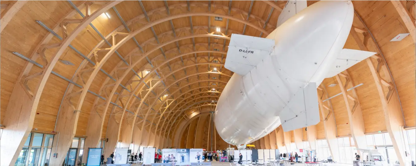 Dieses Foto zeigt die Werbeartikelmesse NRW 2025 im Luftschiffhangar Mülheim an der Ruhr. Zwischen dem imposanten Holzgewölbe und dem riesigen Zeppelin präsentierten zahlreiche Aussteller ihre neuesten Werbeartikel und Branding-Lösungen.